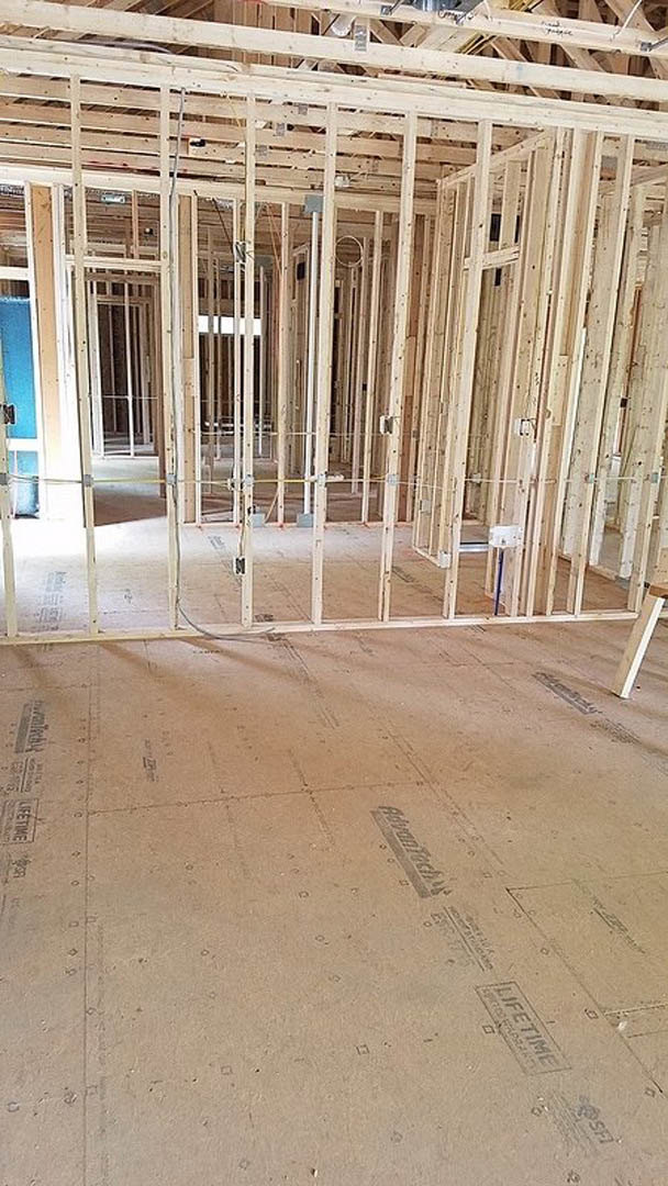 Wood-framed room under construction with exposed beams, subfloor marked with black text, and a white-framed window.