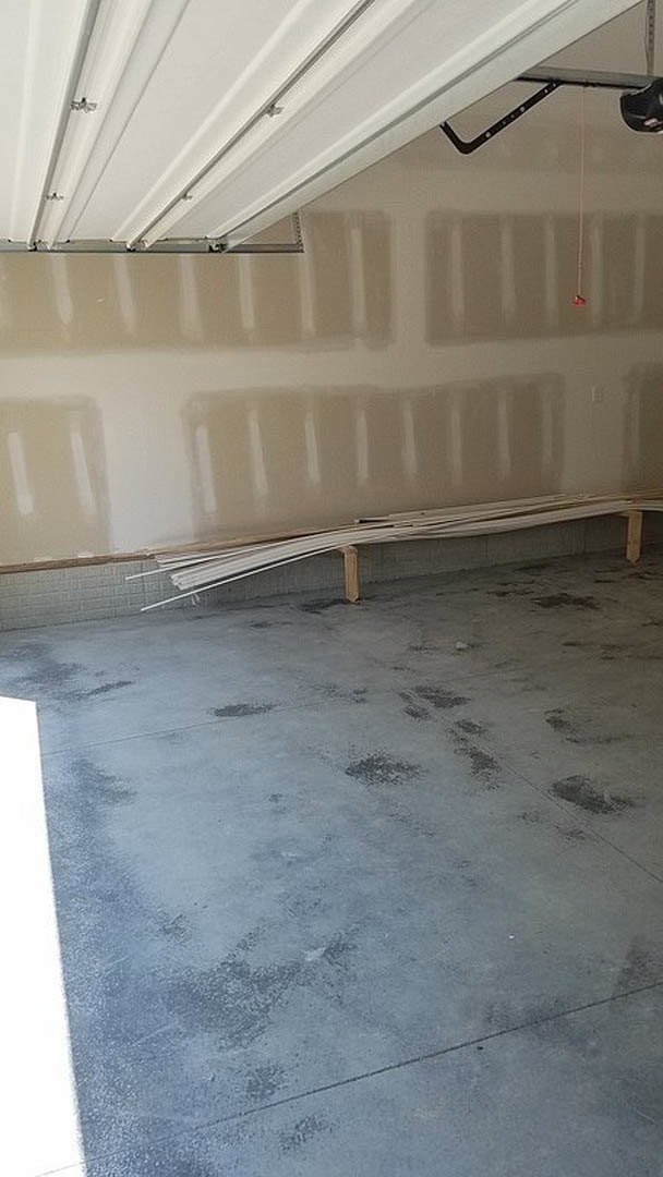 Concrete floor with stacked wood planks, plaster wall, wooden bench, roller blind, and partially painted garage door in a modern interior space.