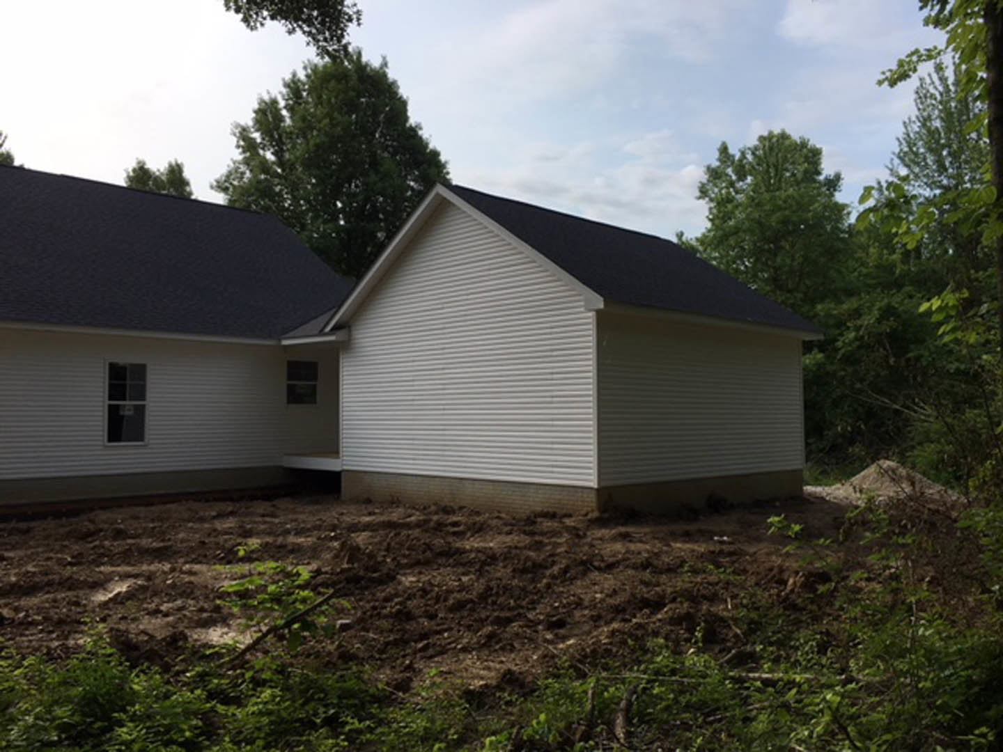 White house with black roof under construction, surrounded by dirt patch and mature trees; white-framed windows with small square panes visible on exterior siding