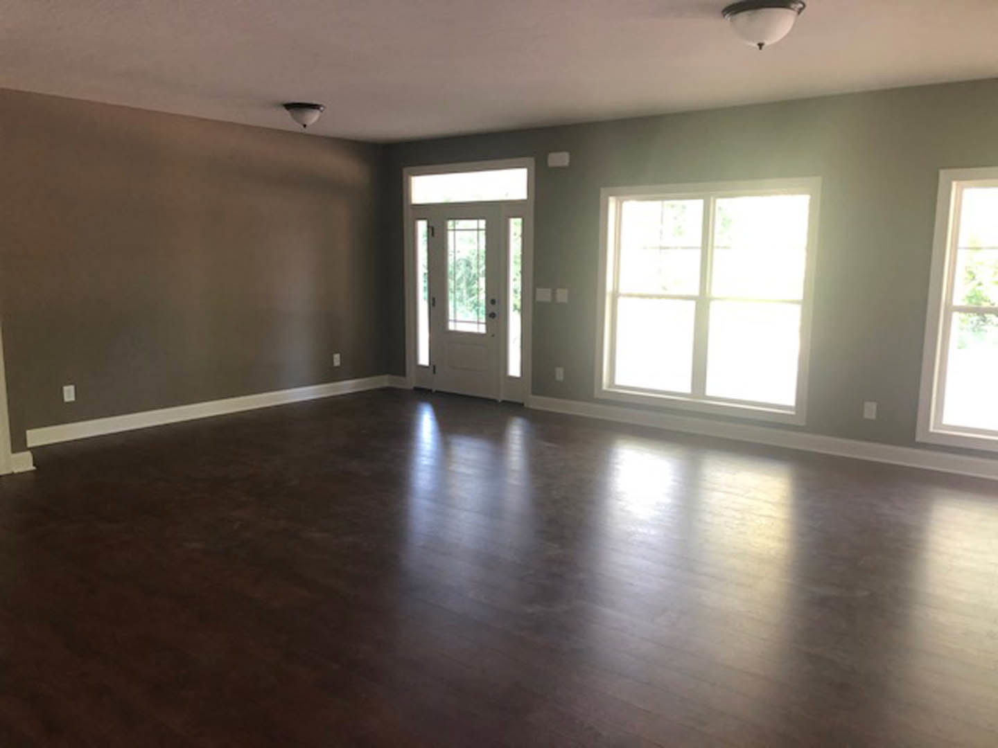 Bright room with wood flooring, white framed windows, and a white door with glass panels; sunlight streams through the windows, highlighting the laminate floor and neutral walls.