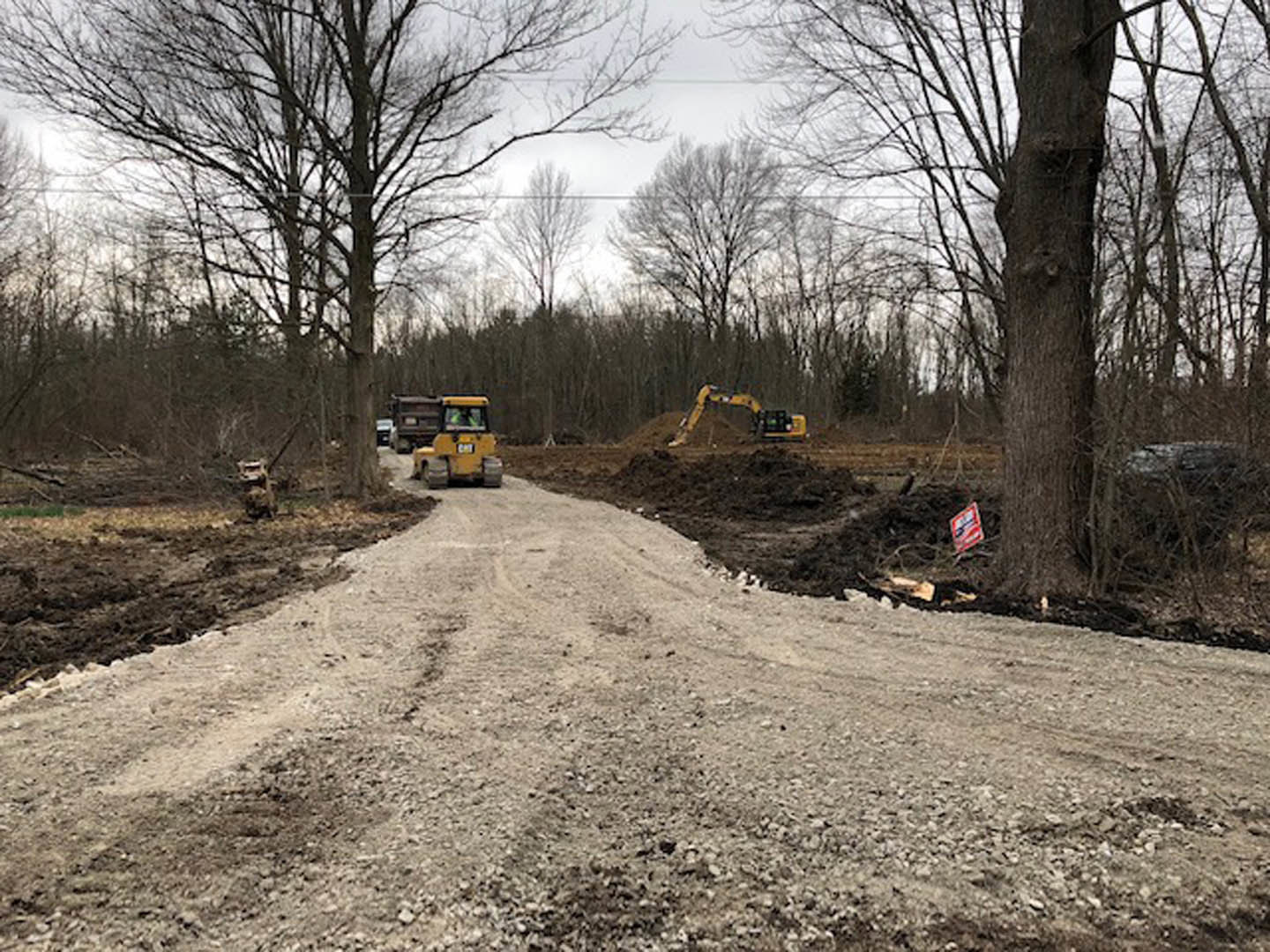 Dirt road lined with trees, yellow construction tractor parked on soil, truck visible in background