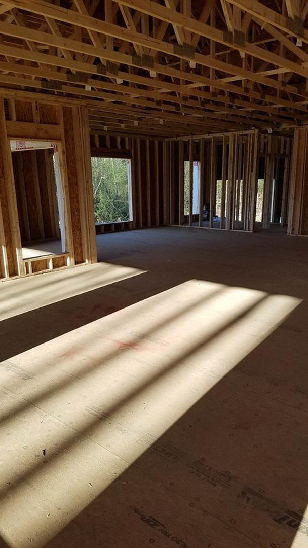 Sunlit room featuring exposed wood framing, large window overlooking green bush, wooden ceiling and wall details, light reflecting on hardwood floor.