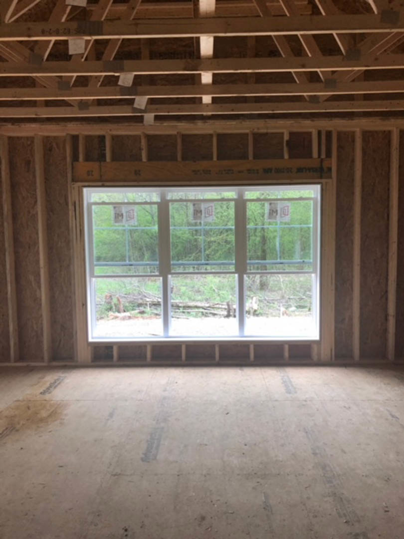 Large window framed by wood beams, concrete floor with dark markings, wooden ceiling detail, view of fence and trees outside