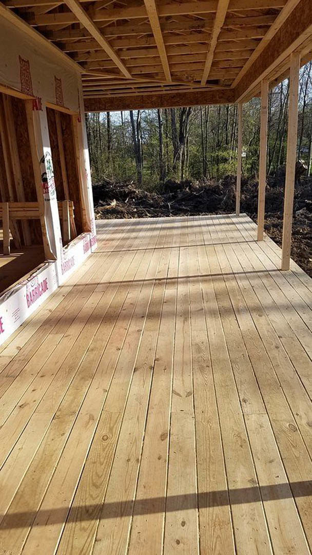 Wood deck with horizontal planks, wooden railings, and a wood-framed structure featuring exposed beams and a sloped roof in the background; sunlight casts shadows across the deck