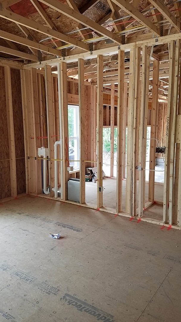 Wood-framed room under construction with exposed beams, unfinished walls, visible window, door opening, and concrete floor