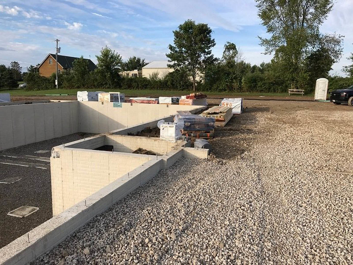 Gravel pile and black truck parked beside partially built white brick house, white door with metal handle, leafy tree in background, rectangular construction materials on ground
