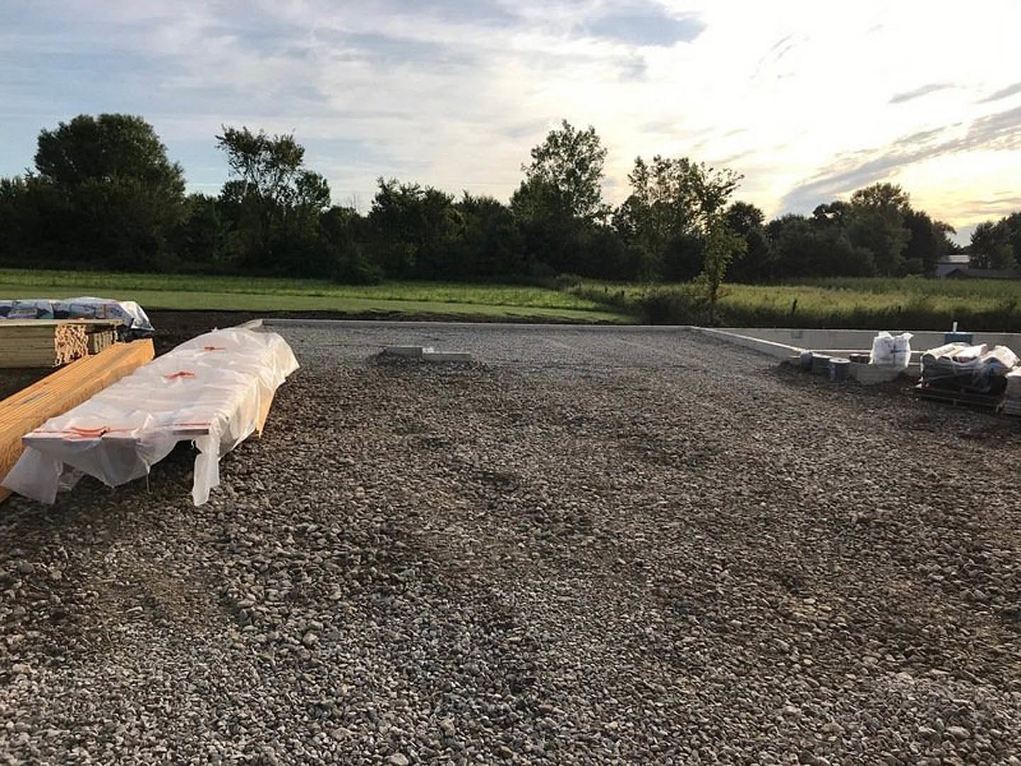Gravel-covered construction site with wooden bench, white plastic sheet on wood, curb edging a gravel road, grassy area, distant trees, and white bag hanging on fence