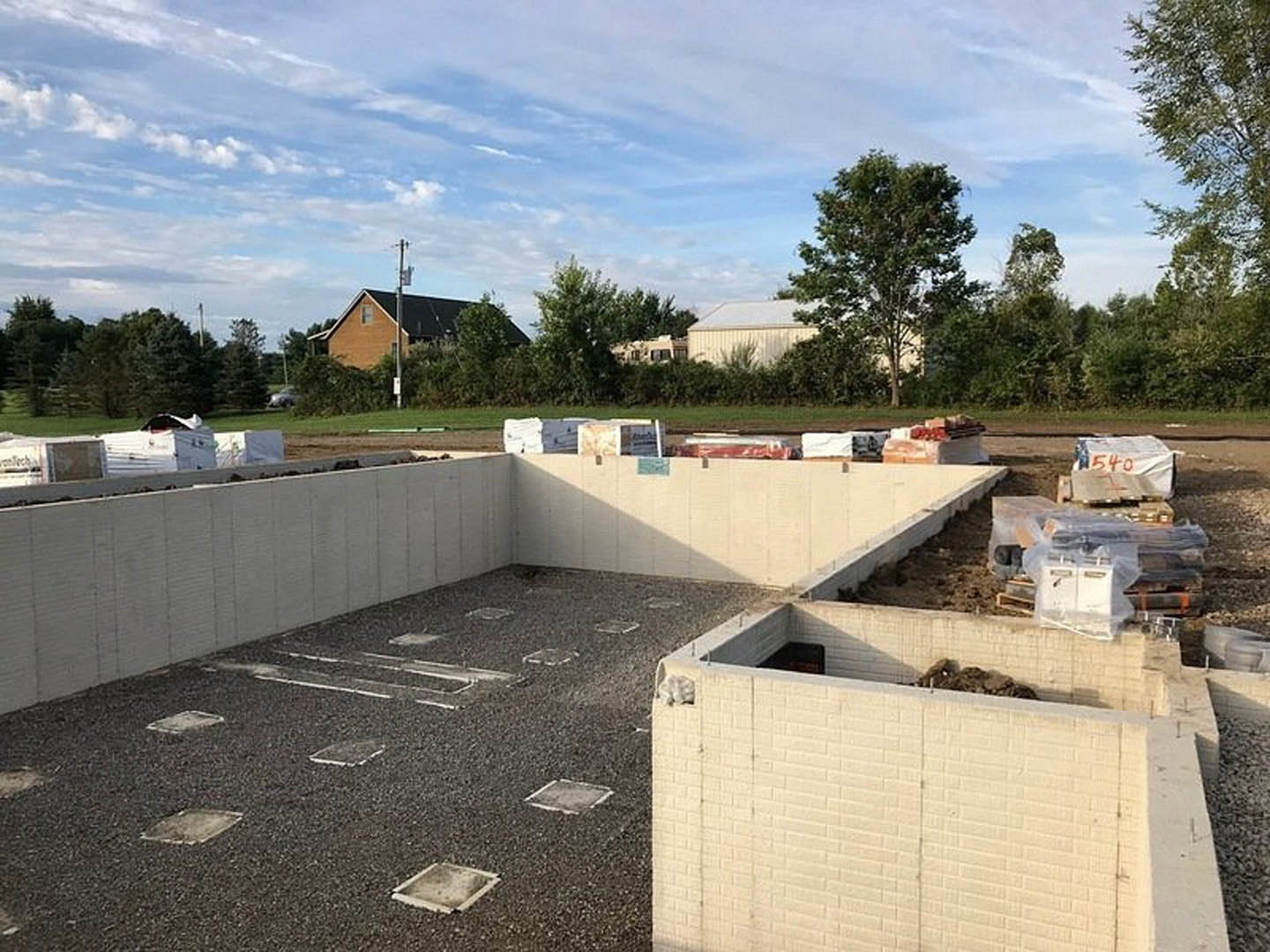 Construction site with stacked cement blocks, scattered materials, partially built wall with exposed dirt, leafy tree, and unfinished house in the background under a cloudy sky