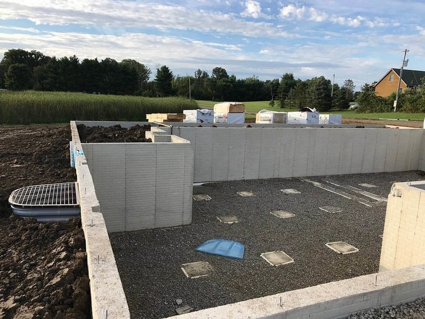 Dirt construction site bordered by trees, square concrete slab and white marker on ground, blue sky with scattered clouds overhead