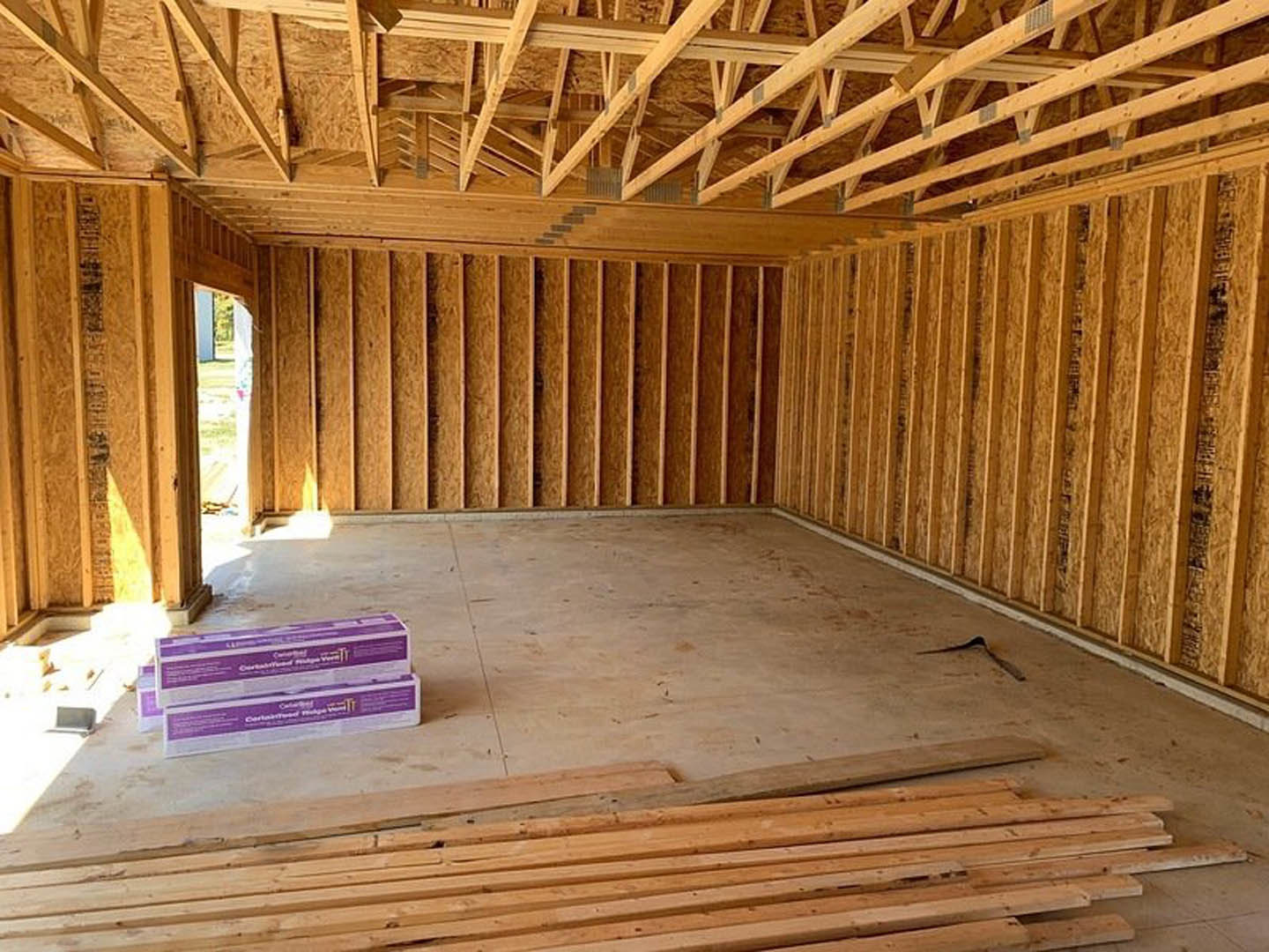 Living room with exposed wood ceiling beams, stacked lumber against one wall, hardwood flooring, and unfinished walls.