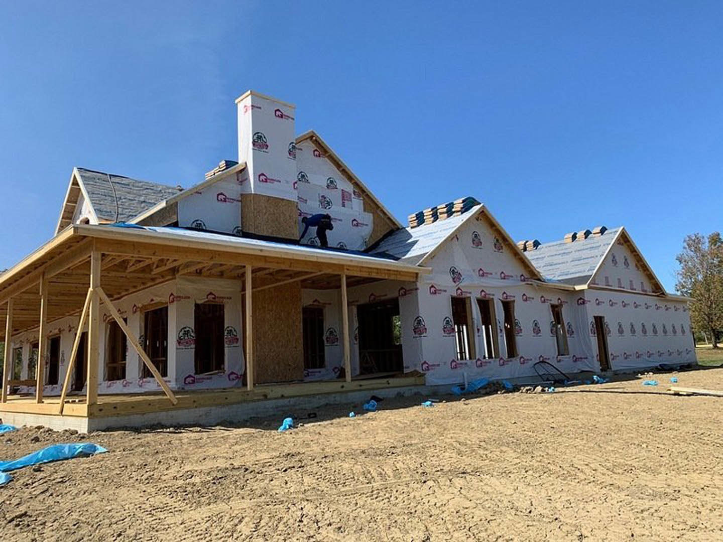 Wood-framed house under construction with a worker on the roof, blue tarp and bags scattered on dirt ground, leafy tree nearby