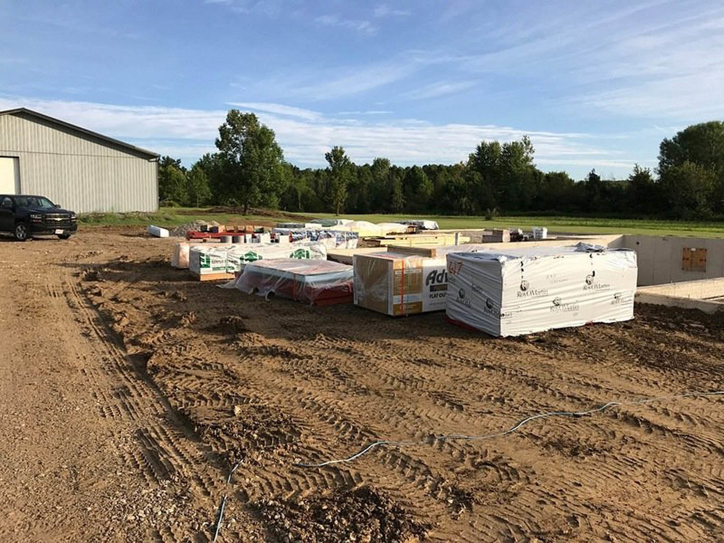 Stacked construction materials including large white boxes wrapped in plastic, black truck and car parked on dirt field, grassy area and trees in background under partly cloudy sky