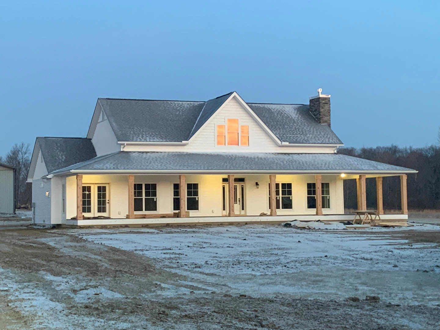 White two-story house with black-framed windows, expansive covered porch, snow-covered lawn, and wooden fence