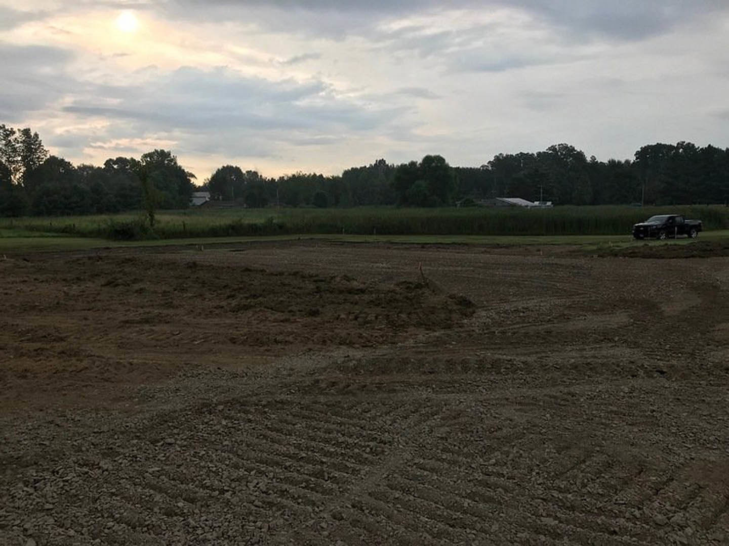 Dirt field with tire tracks, black van on road, tractor in background, cloudy sky, bright sun, group of trees along edge of rural landscape