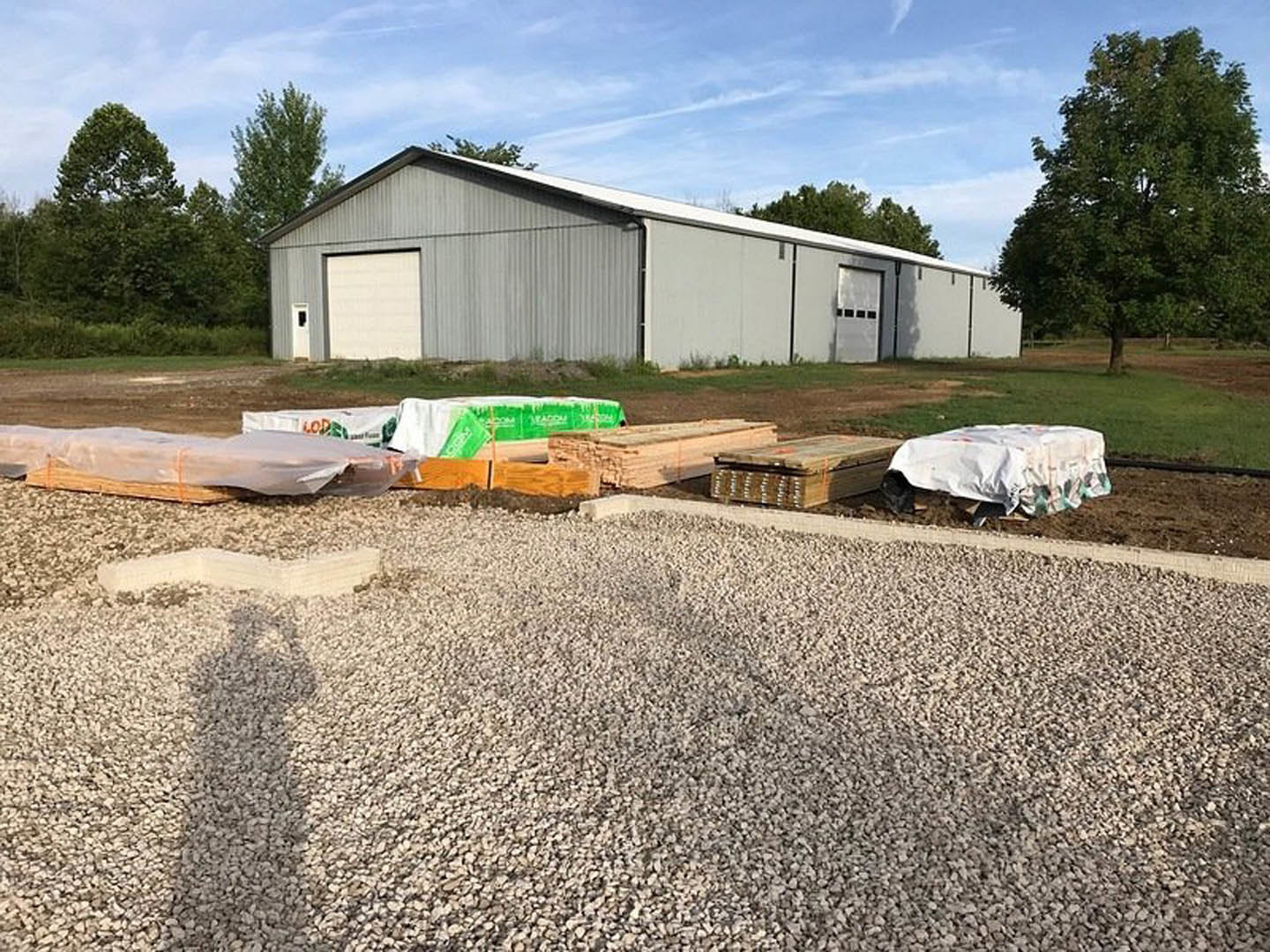 Grey house with white roof surrounded by pallets and gravel, leafy tree nearby, white tarp spread on ground, construction materials scattered around, close-up window visible.