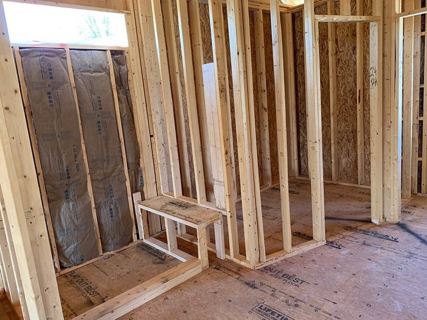 Room under construction with exposed wood framing, plywood walls, wooden shelf, white rectangular object illuminated in background, and wooden frame with black text resting on