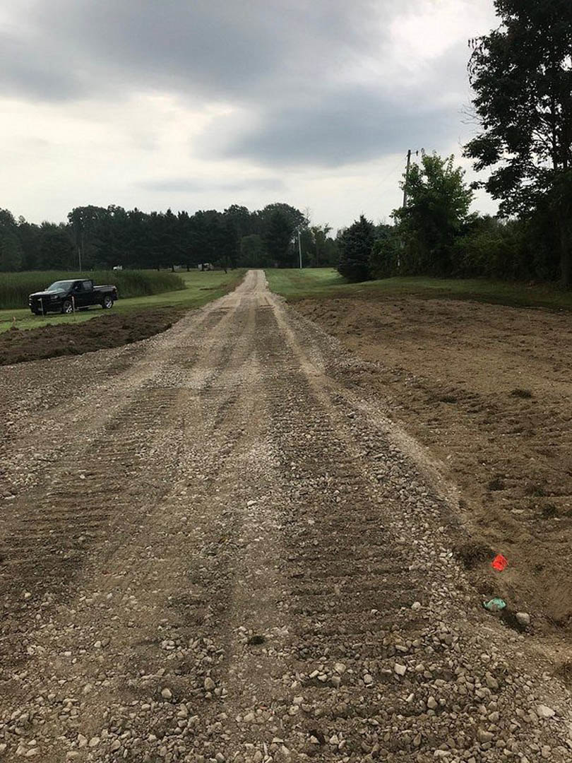 Dirt road with deep tire tracks leading to a black truck parked on grassy ground, surrounded by scattered trees under a cloudy sky