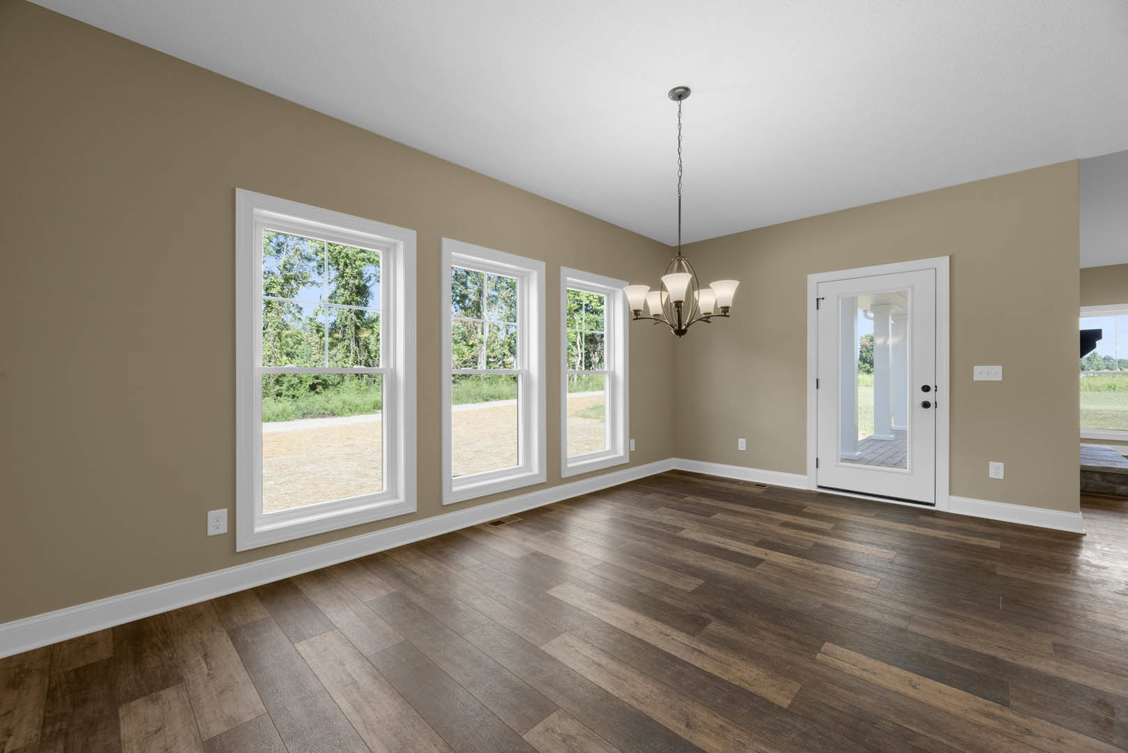 Chandelier hanging from white ceiling, large windows with tree views, wood flooring, white door with black handles