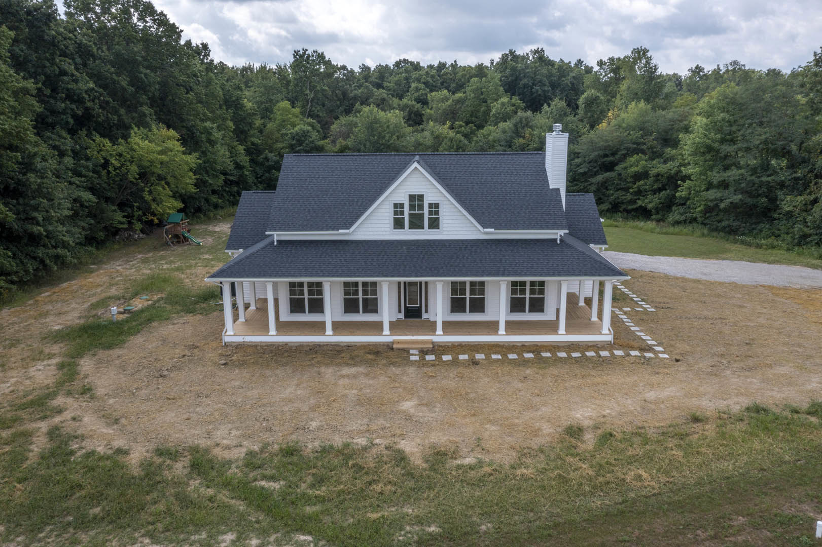 Two-story house with gray siding and white trim, spacious concrete driveway, covered front porch, grassy yard bordered by mature trees, cloudy sky overhead