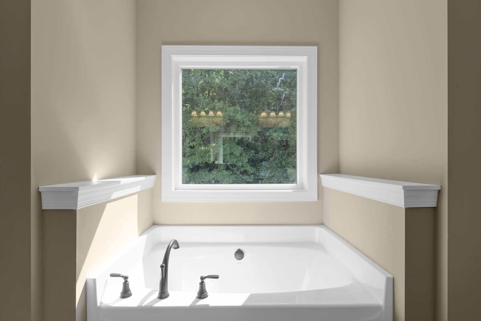 Freestanding white bathtub beneath large window with trees visible outside, chrome faucet, white shelf illuminated by natural light, detailed white molding, wall-mounted hook