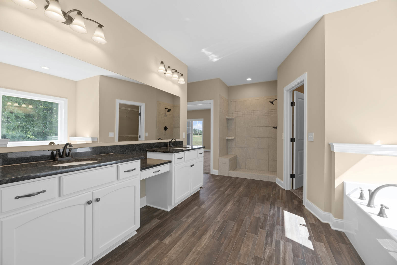 Bathroom with wood flooring, white door and frame, window showing trees outside, close-up of chrome bathtub faucet, wall-mounted light fixture, shadow of person cast on wall