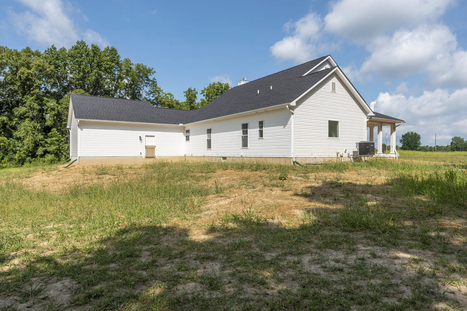 White farmhouse with black roof, surrounded by grassy yard, brick wall, and trees under partly cloudy sky