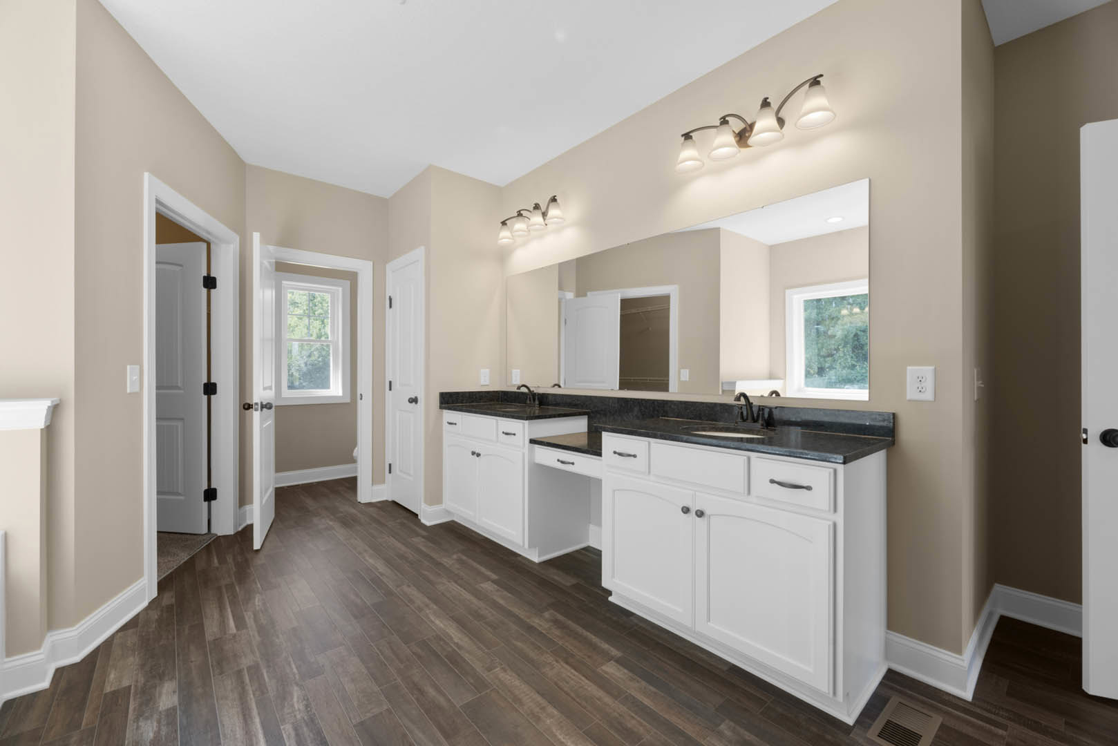 Bathroom with expansive mirror above white cabinetry, quartz countertop, undermount sink, multi-bulb light fixture, white electrical outlet, window with white frame, and door