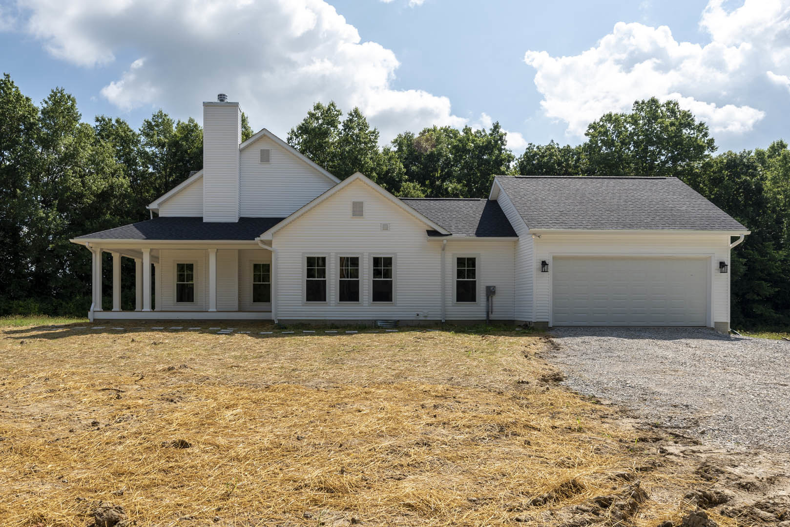 White siding house with black roof, expansive concrete driveway, manicured lawn, mature trees in background, blue sky with scattered clouds