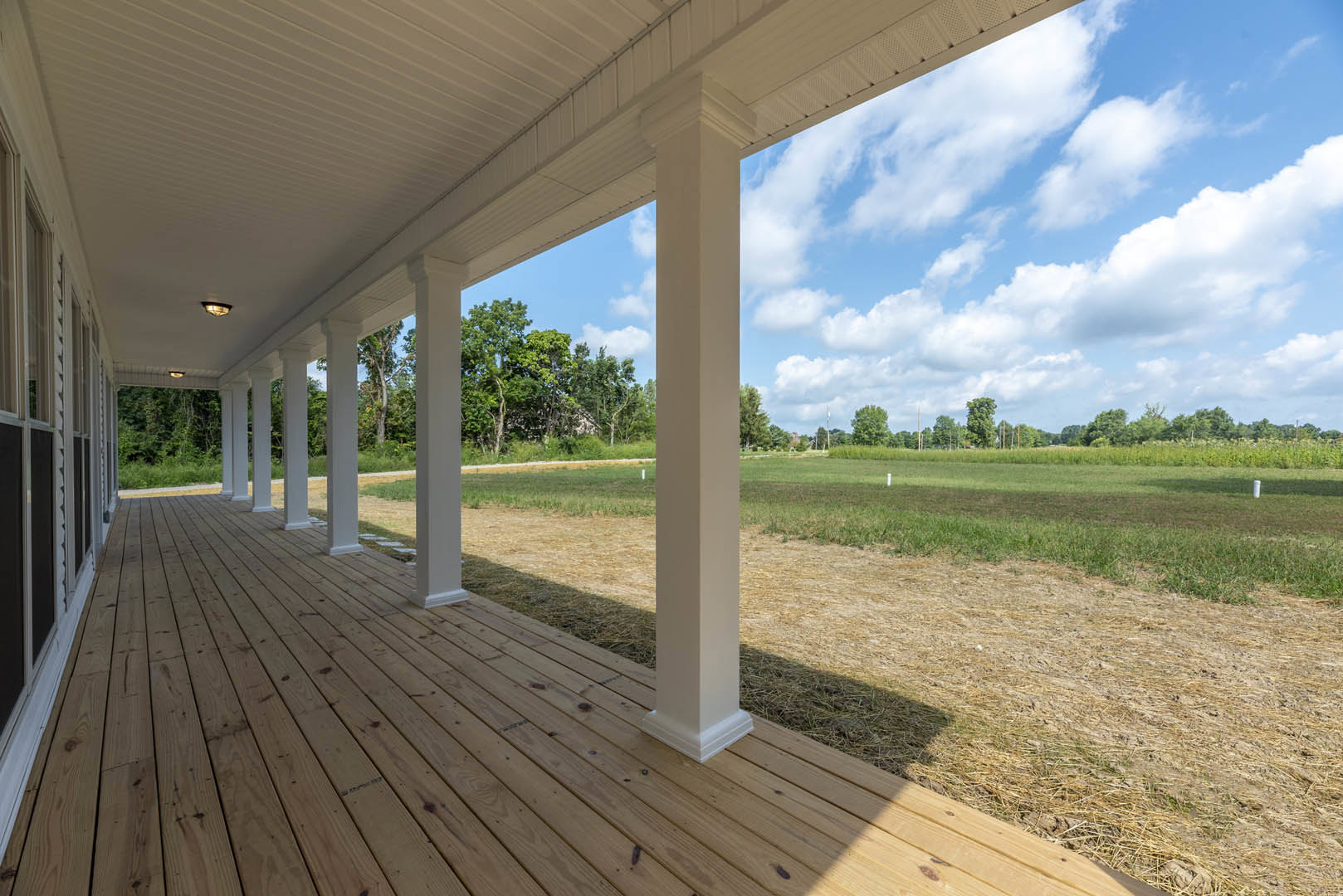 White porch with wooden pillars, shaded roof, expansive green lawn, mature tree with leafy branches, blue sky and clouds in background