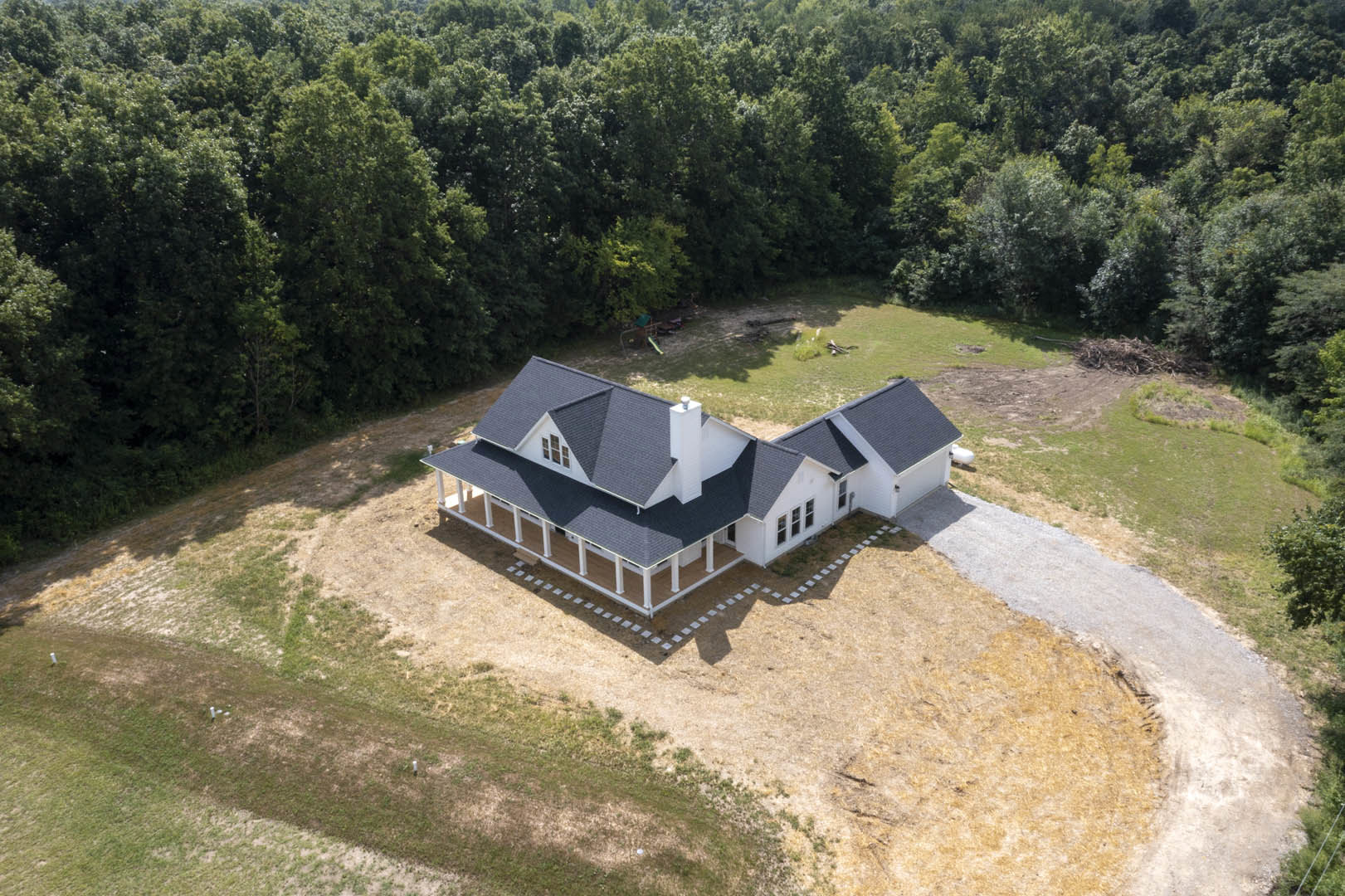White house with black roof, expansive driveway, large front porch, grassy yard bordered by mature trees