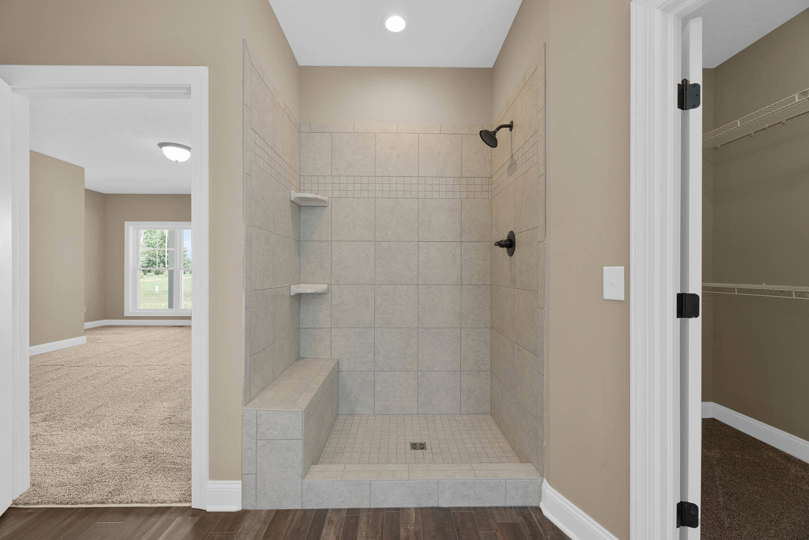 Walk-in shower with black faucet on white tile wall, window overlooking grassy field, brown-framed white bench, black soap dispenser on countertop, carpeted area adjacent to white