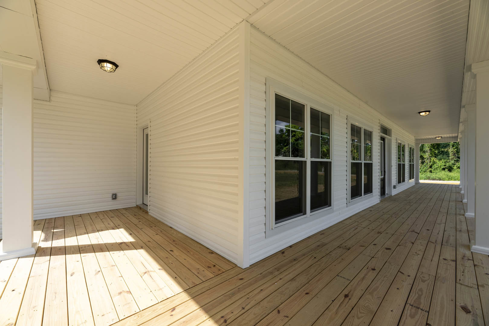 White exterior with black trim, wood flooring inside, ceiling light fixture, wooden deck, large windows framing forest views