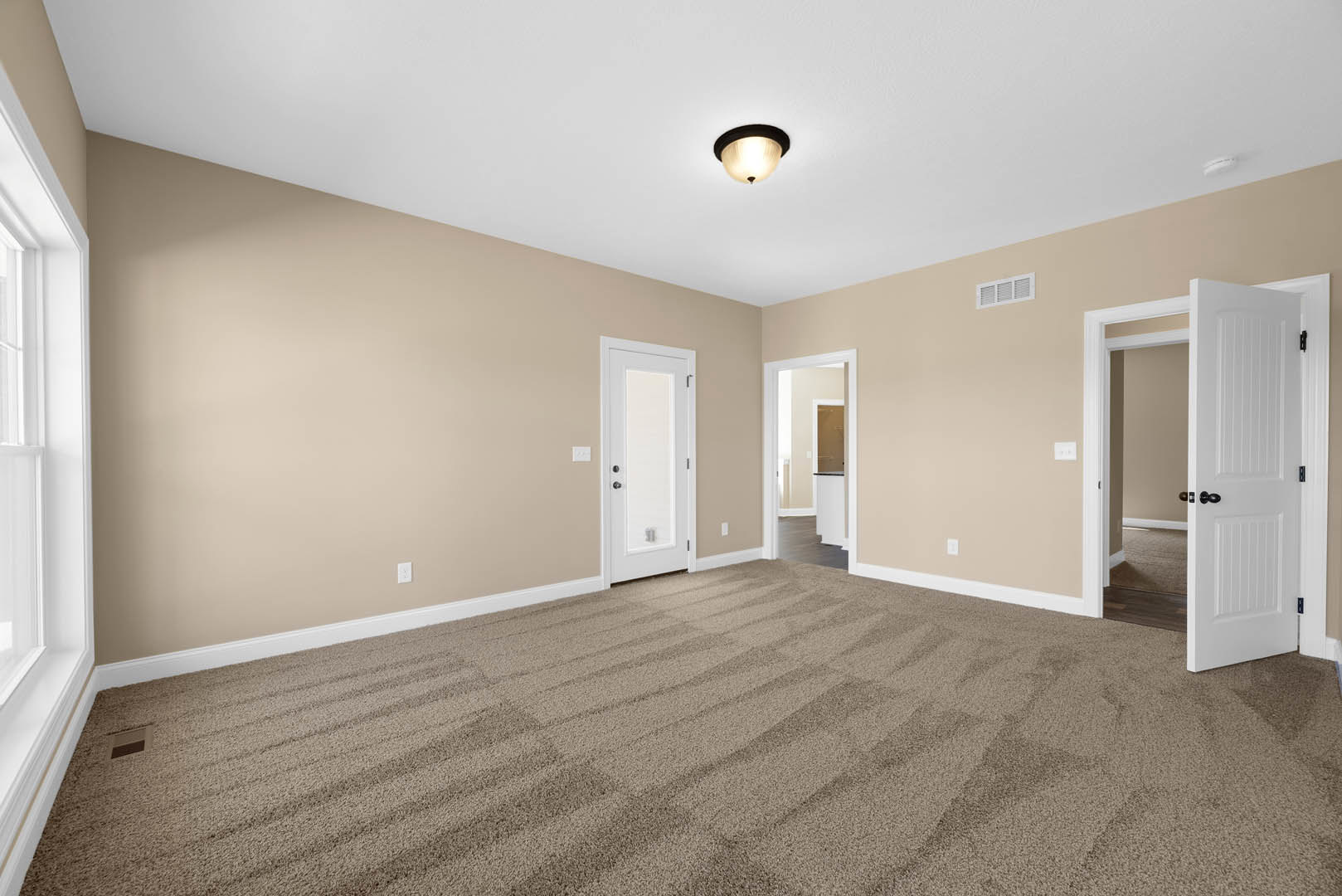 Beige walls and carpeted floor, white door with black handle, ceiling light fixture, open hallway with white door and black knobs, simple molding along walls and ceiling