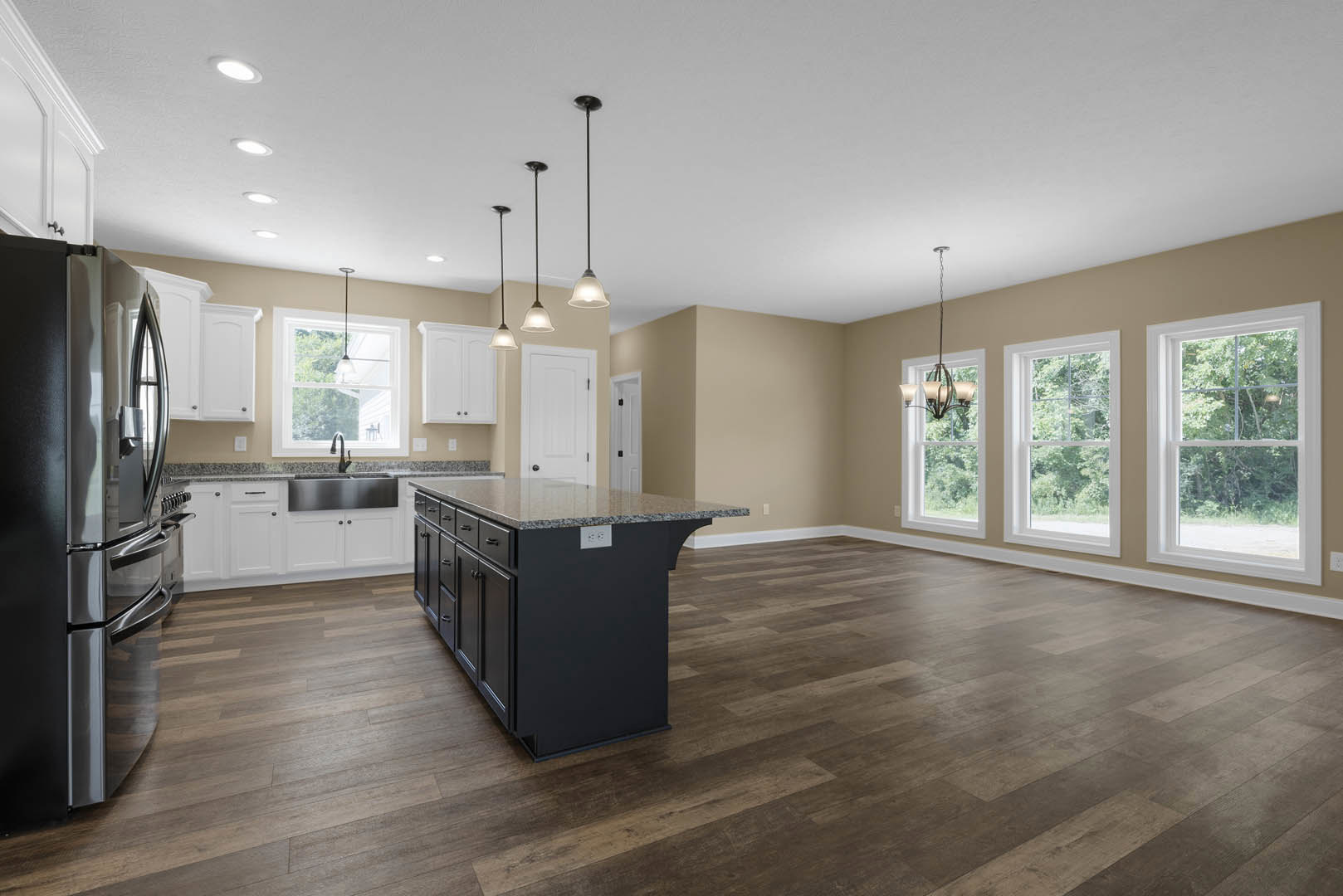 Kitchen with wood flooring, black island featuring granite countertops, stainless steel refrigerator, window overlooking trees, sink beneath pendant light fixture, white cabinetry