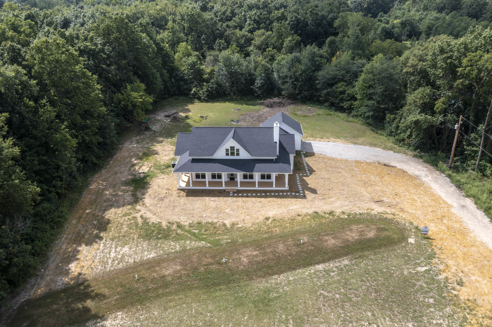Two-story custom home with gray siding and large front porch, nestled among mature trees, set on a grassy lot with a dirt driveway and mountain views in the background