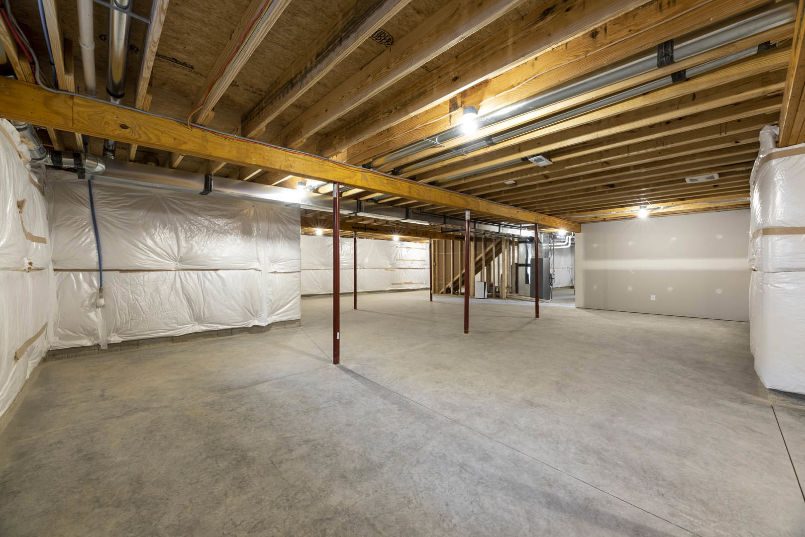 Exposed wooden ceiling beams, wood-paneled ceiling, concrete floor with red support poles, light illuminating a wall, stack of toilet paper wrapped in plastic