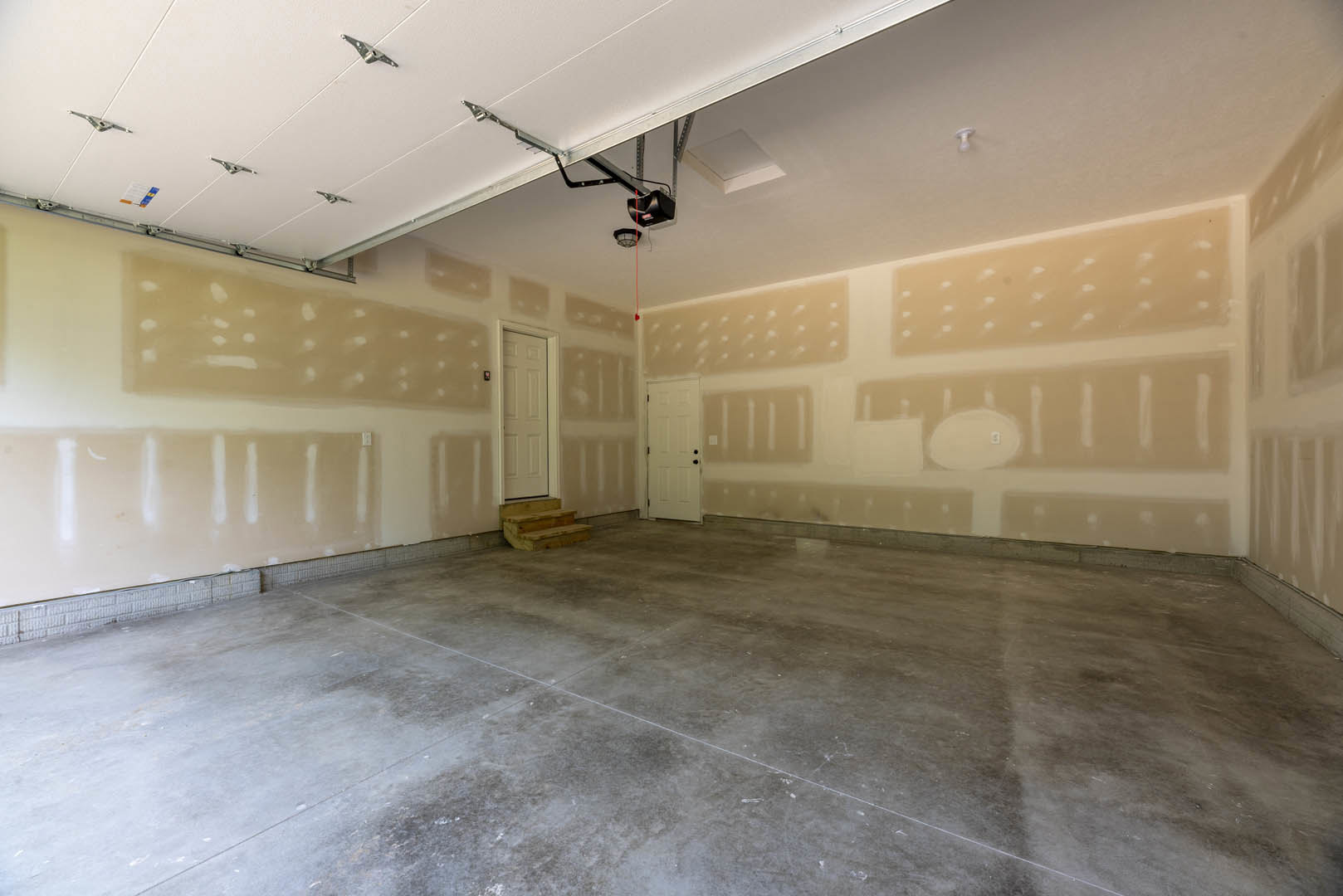 Garage interior with smooth concrete floor, white paneled door featuring black hardware, adjacent staircase with white risers, and walls finished in light plaster.