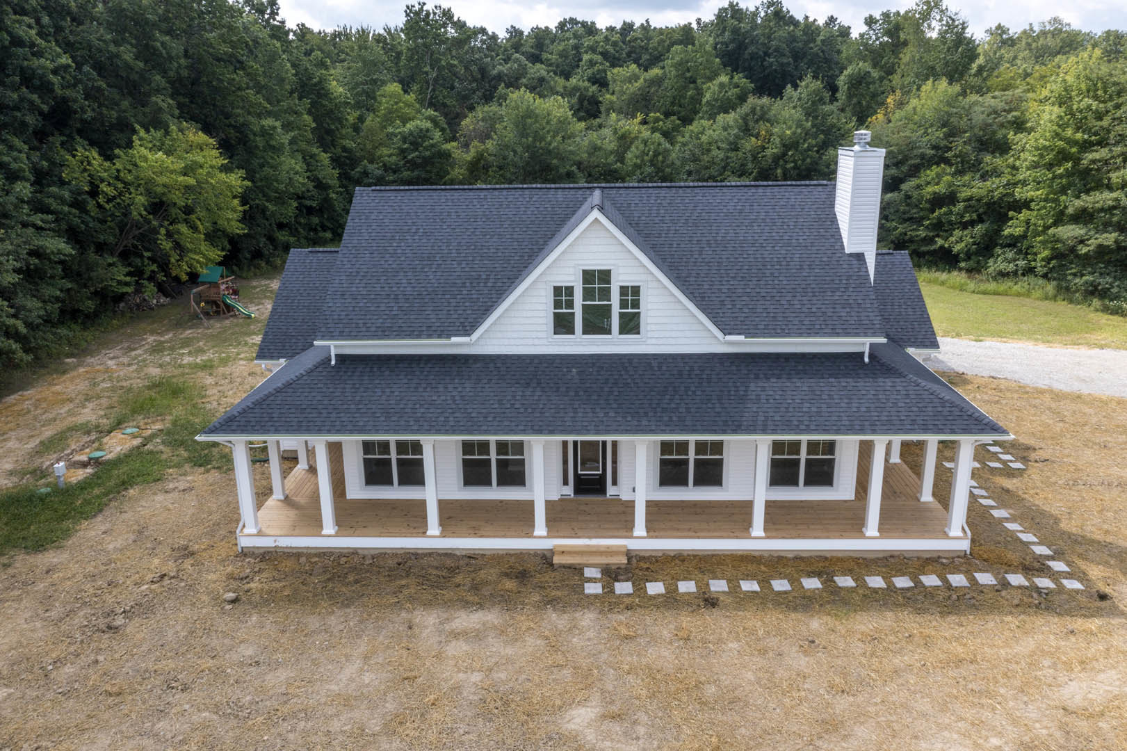 White cottage-style home with black roof, expansive covered porch, white-framed windows, mature trees, and playground slide in the yard