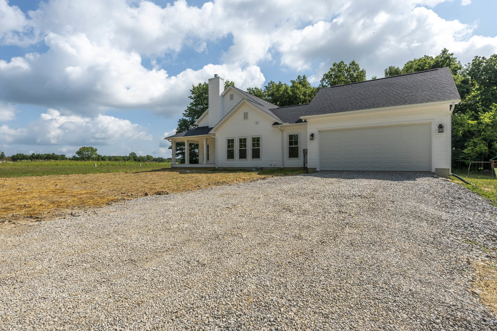 White two-story house with attached garage, gravel driveway, green lawn, and mature trees in the background under a partly cloudy sky