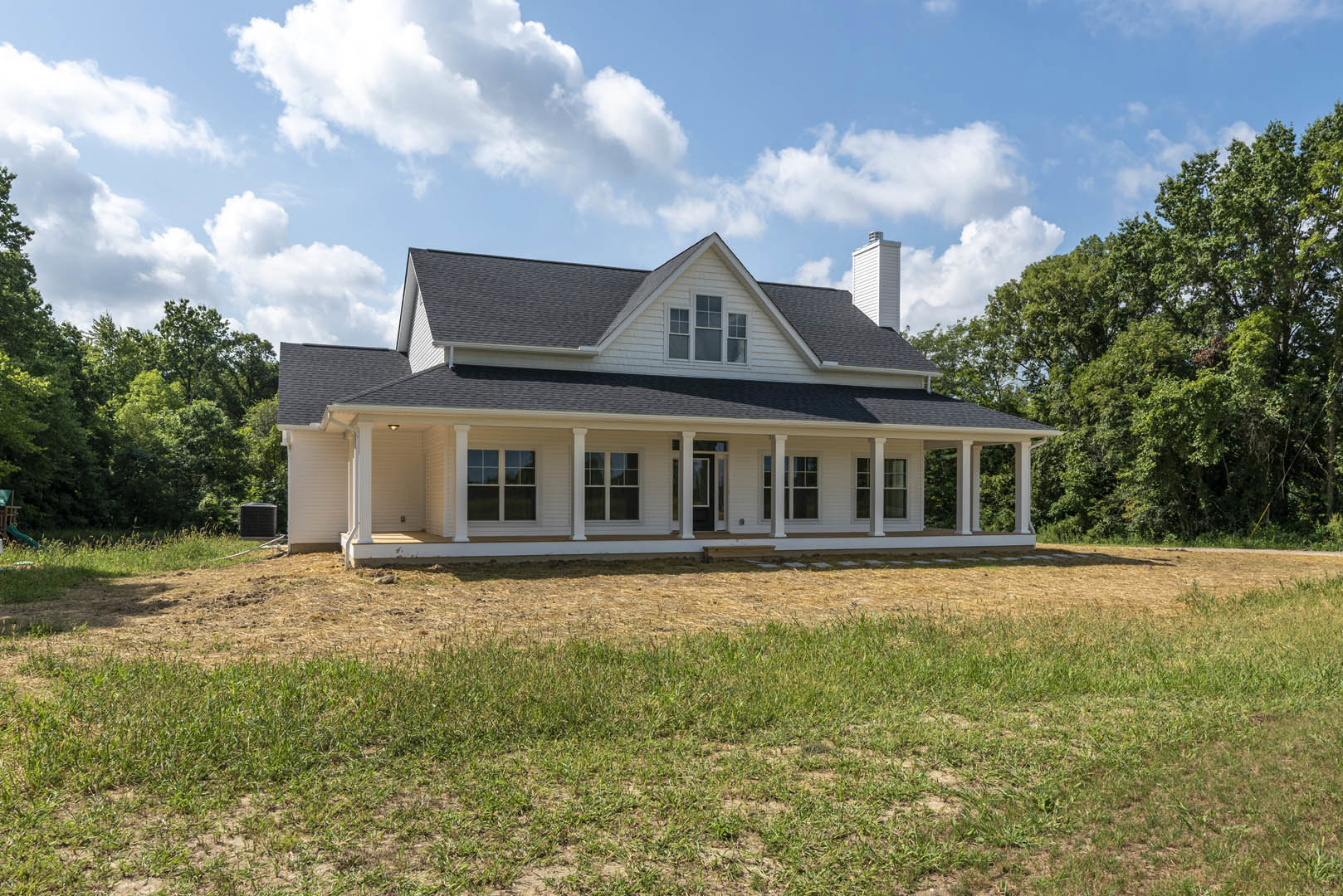 White two-story house with wide front porch, black door, multiple windows, expansive green lawn bordered by a fence, trees in the background, and blue sky with scattered clouds