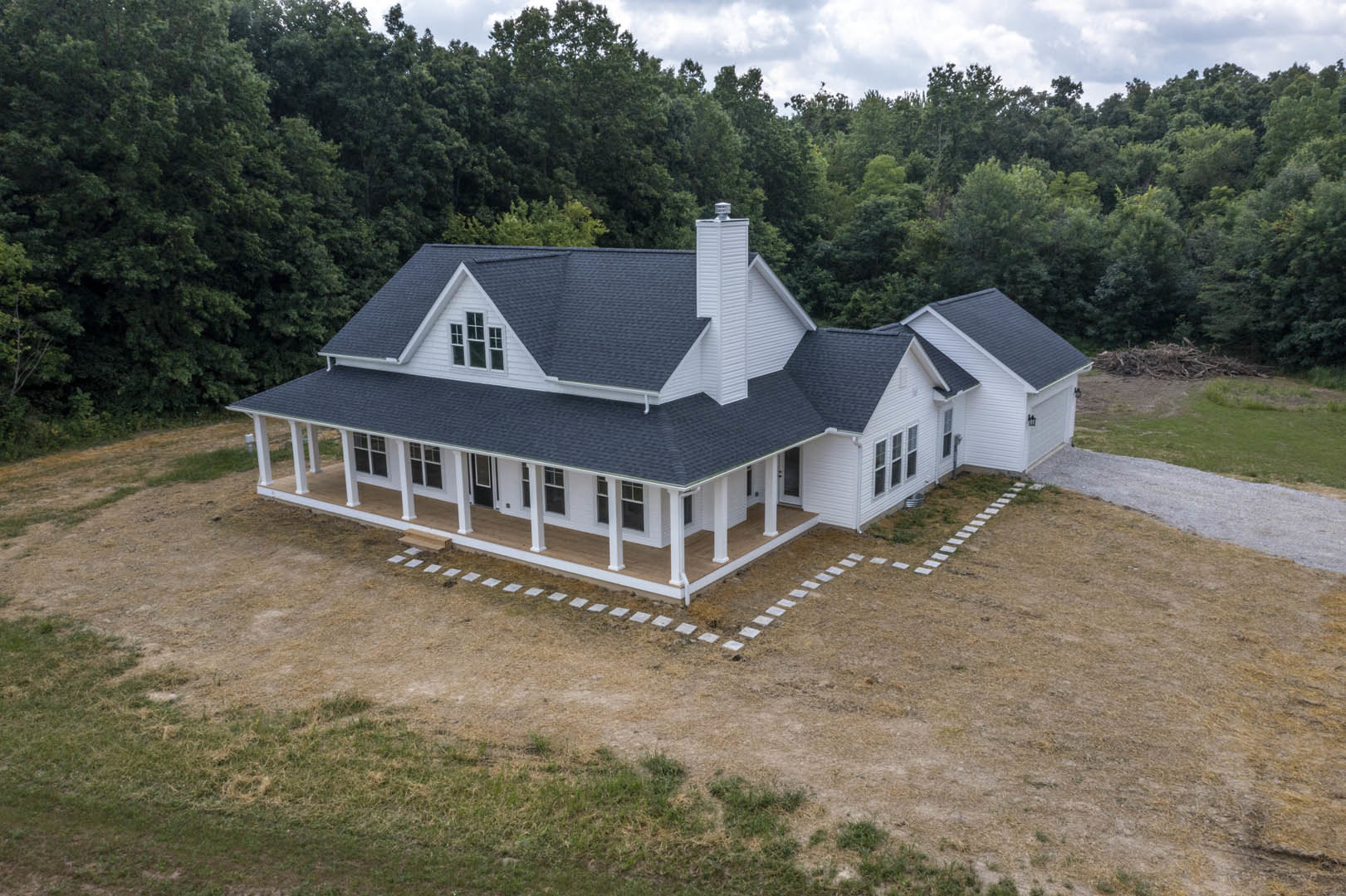 Two-story house with black roof, expansive front porch, white siding, manicured lawn, brick chimney, and mature trees in the background under a partly cloudy sky