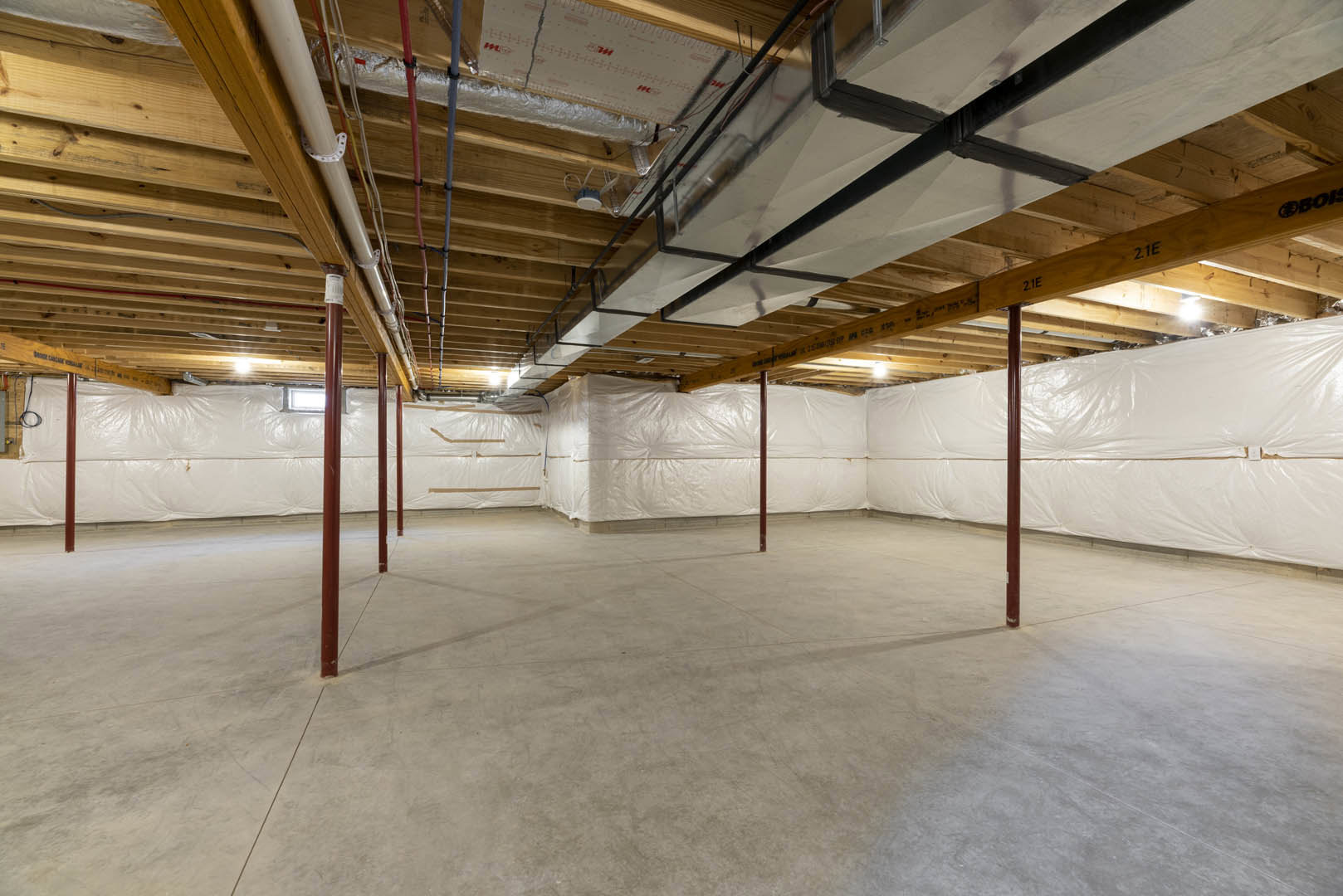 White-walled room with exposed wooden ceiling beams, concrete floor, visible pipes and ducts, brown support pole, and large white plastic coverings in basement space