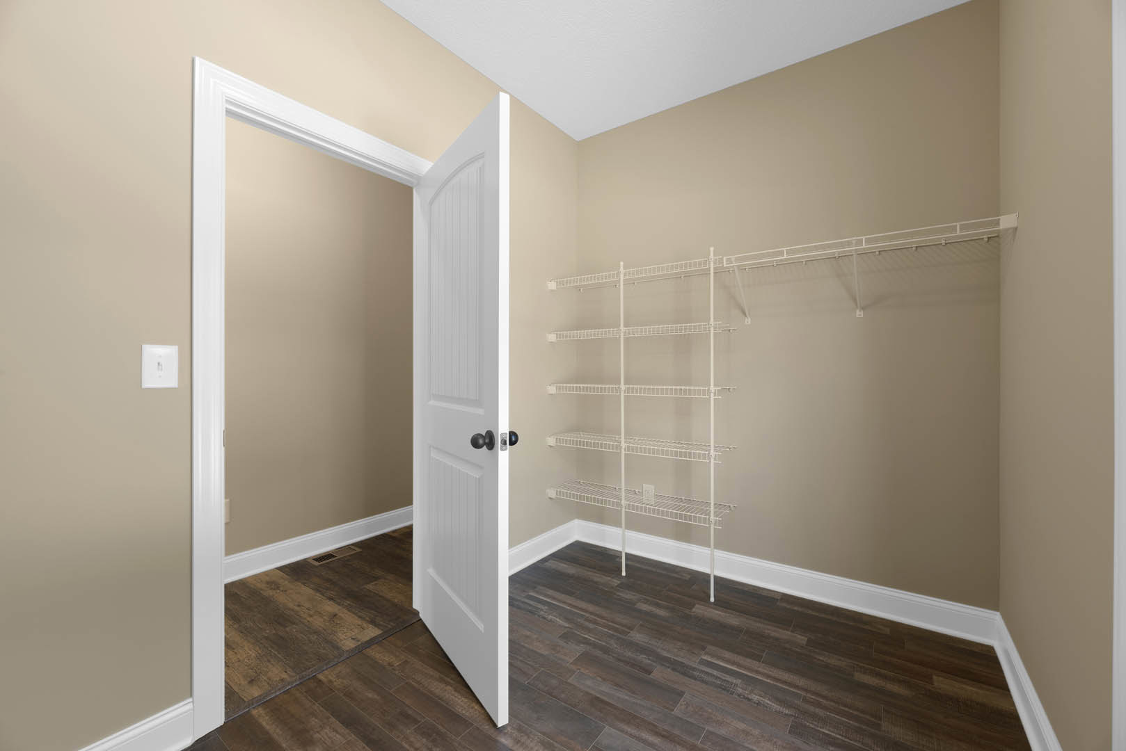 White wire shelving unit against a light-colored wall, open white door with silver hardware, wood flooring with white baseboard trim, light switch near doorway