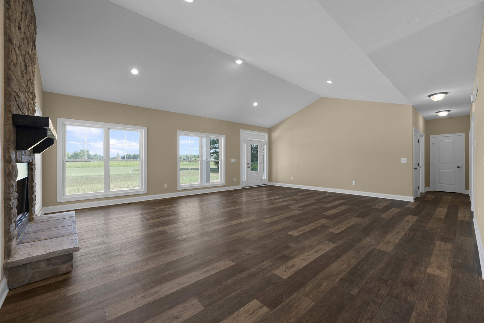 Spacious living area with light wood flooring, stone fireplace, white door with black handle, large windows overlooking a grassy field, and smooth plaster walls