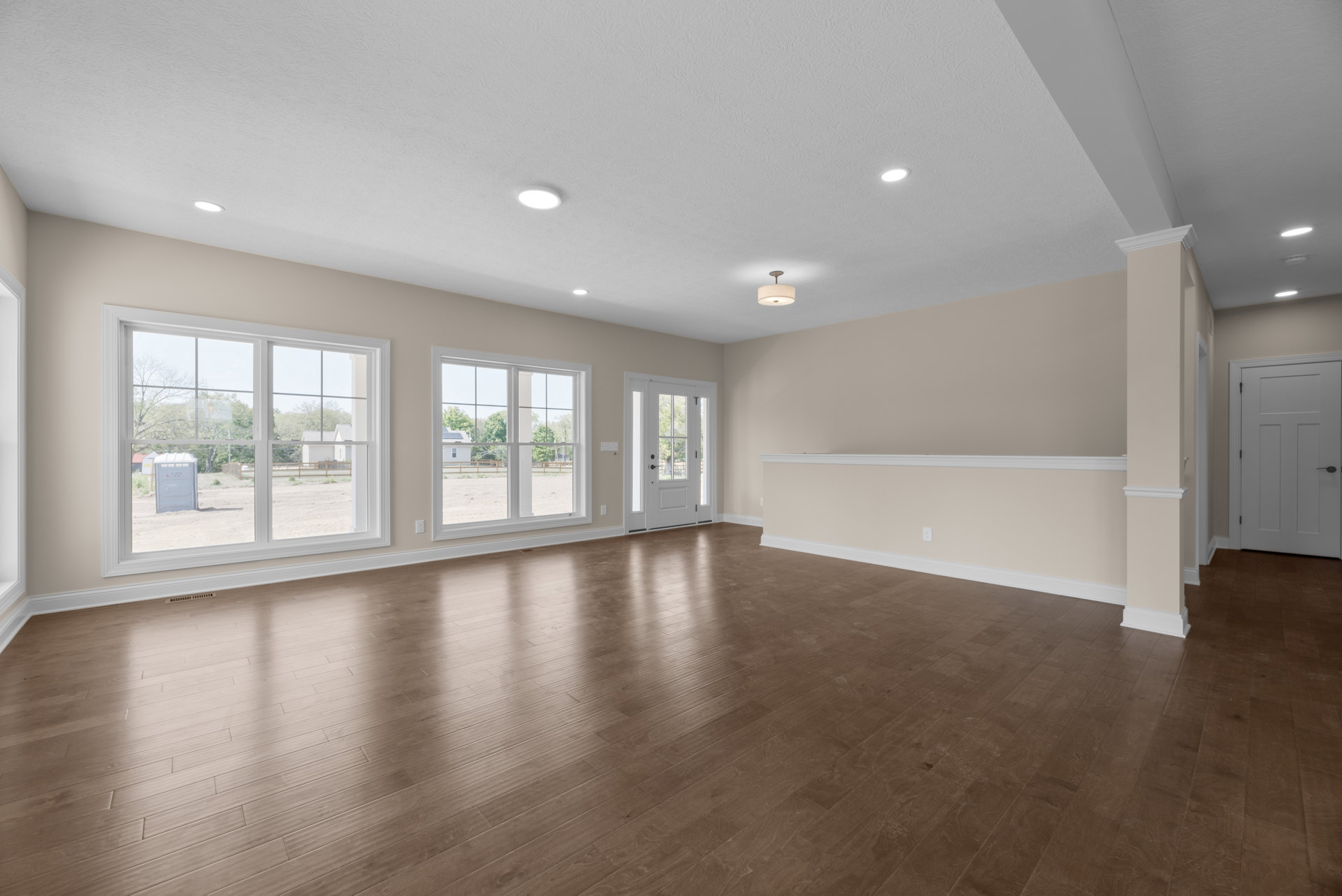 Hardwood floor in a bright room with white walls, large windows, and a white door with black handle and glass panes; view outside shows dirt yard and neighboring house.