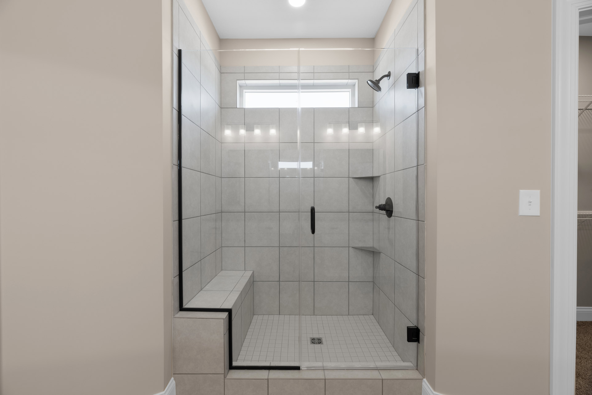 Walk-in shower with frameless glass door, large tile walls, chrome shower head, built-in drain, window for natural light, and nearby light switch on painted wall