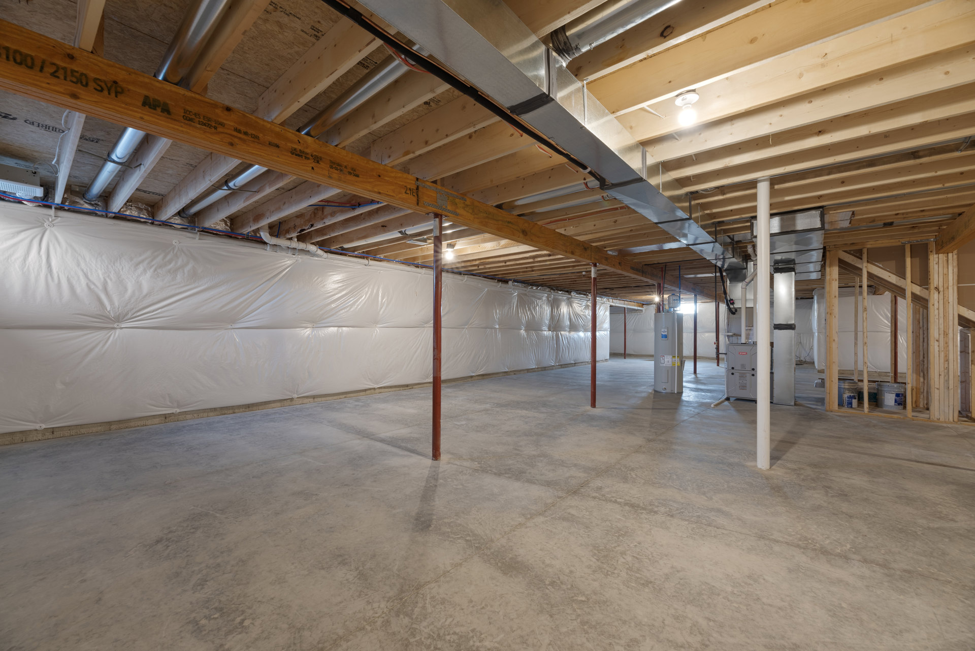 Basement room with white plastic sheet draped over wall, exposed wooden ceiling beams, concrete floor, red steel support poles, and cylindrical container.