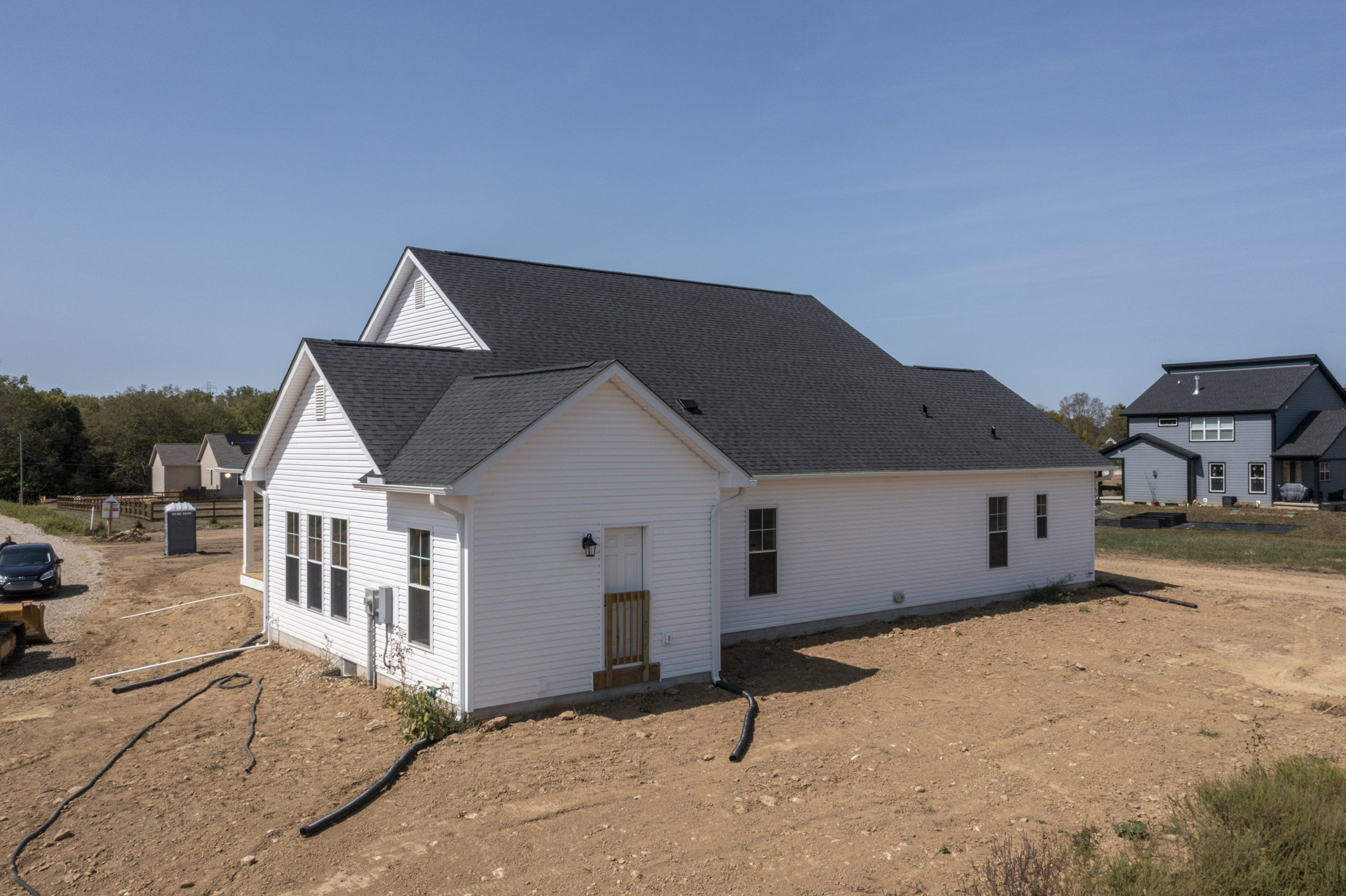 White siding house with black shingle roof, skylight, wooden porch railing, green lawn, black car parked on dirt driveway, man standing beside car
