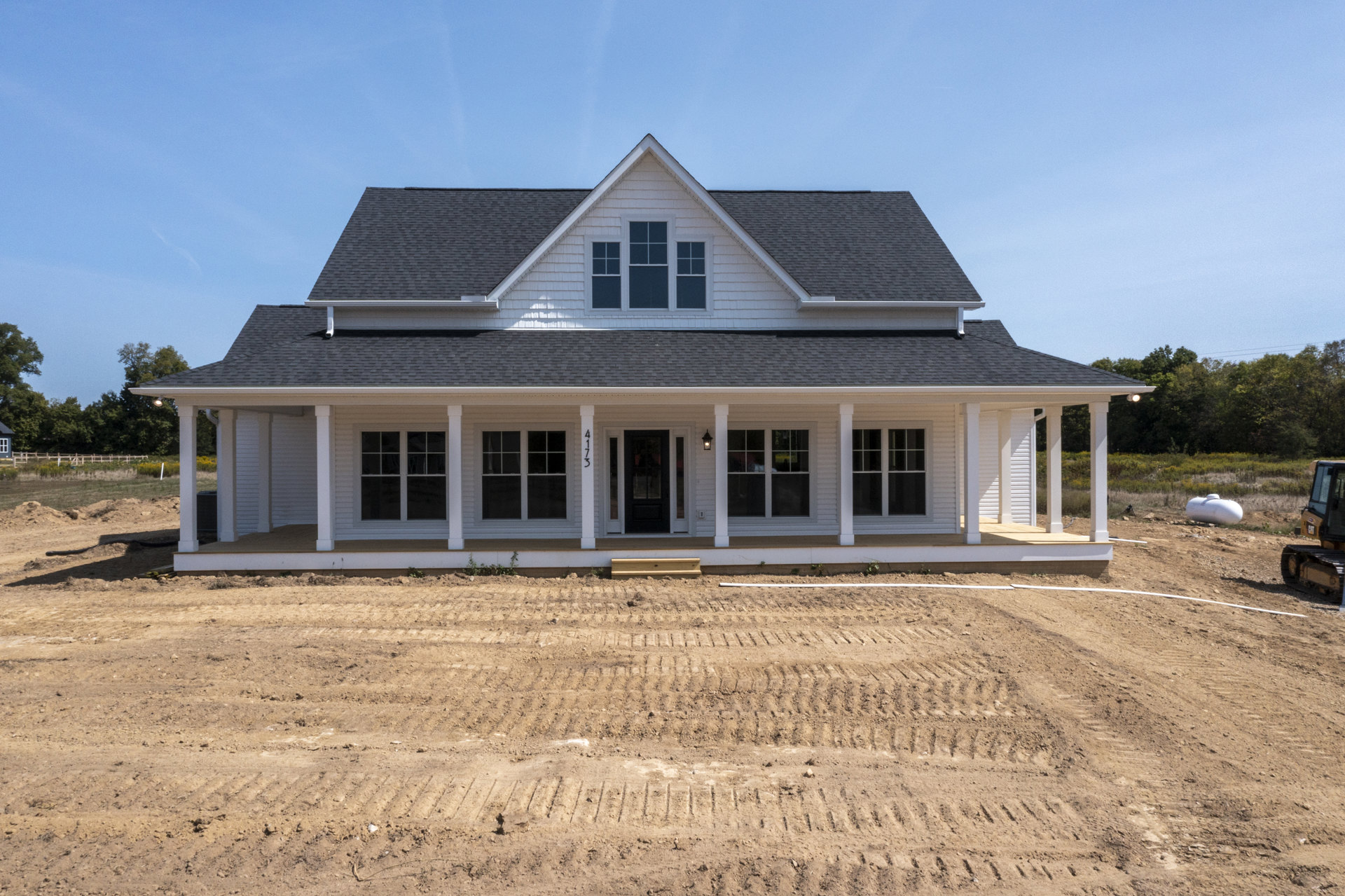 Two-story house with gray siding, black door with glass panels, covered porch, dormer window on roof, dirt field in front, and trees in background