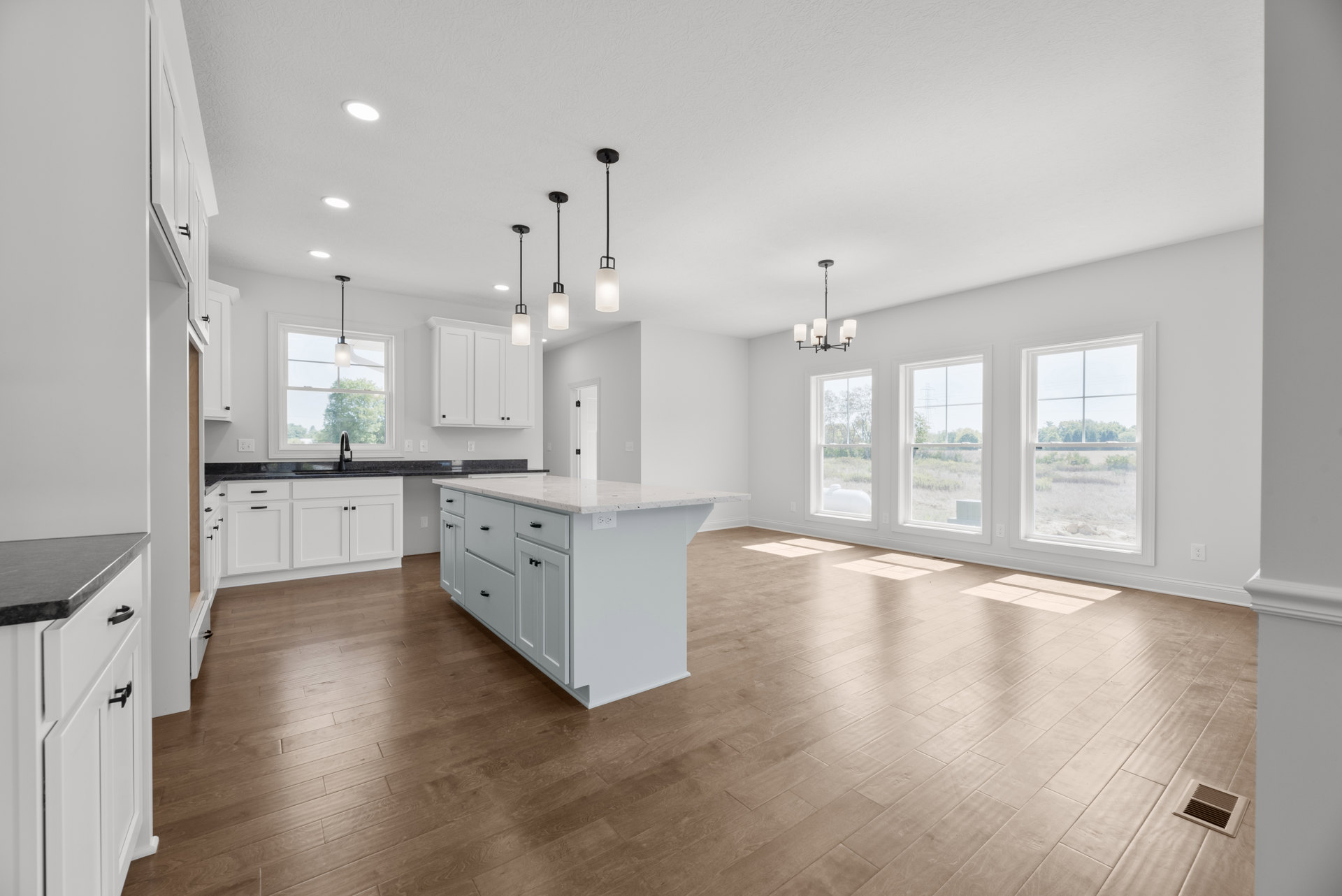 Spacious white kitchen with hardwood flooring, central white island featuring black handles, white cabinetry, ceiling light above sink, and window overlooking open field